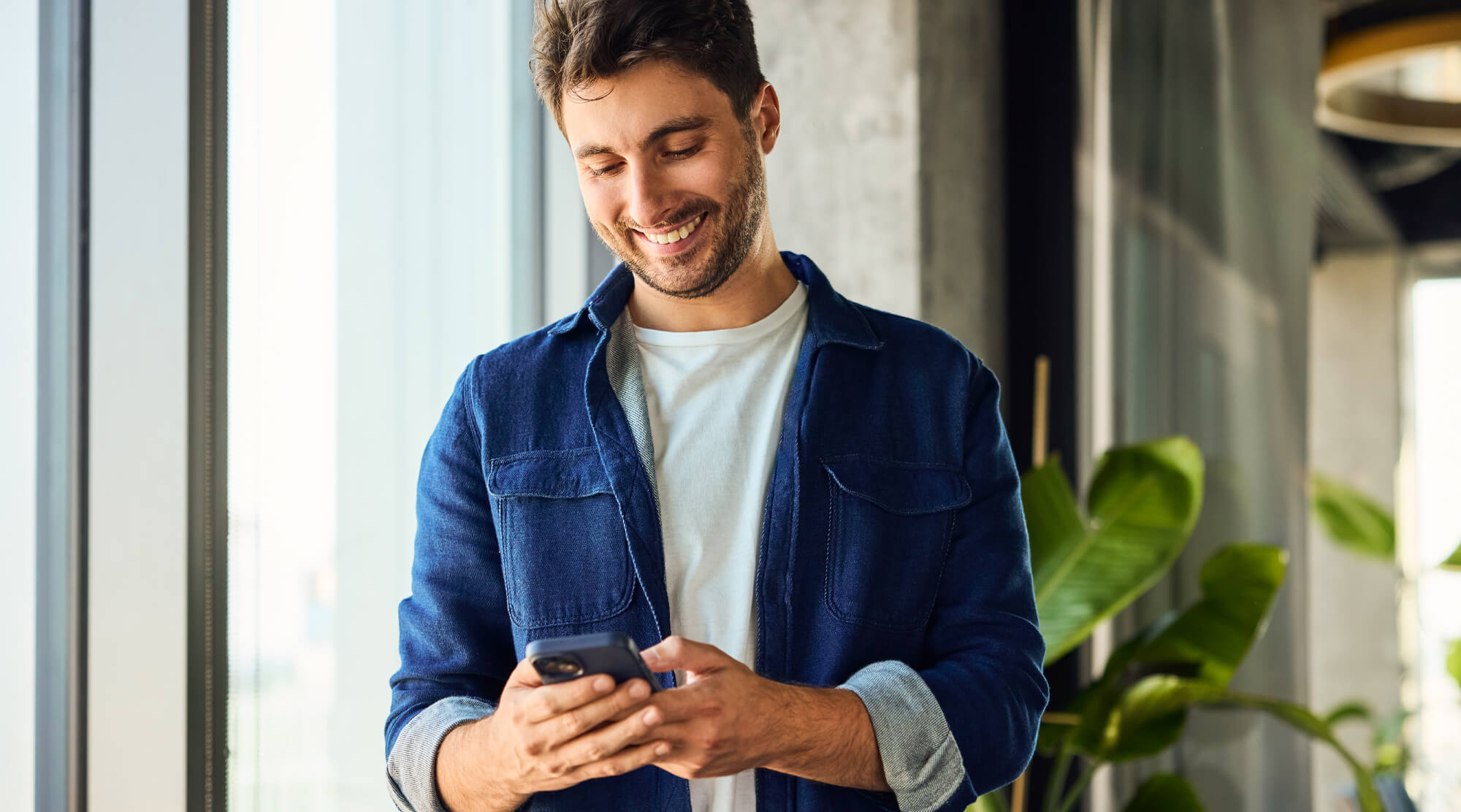 Person in legerer Kleidung steht am Fenster und nutzt ein Smartphone, umgeben von modernen Büro- oder Wohnraumelementen und Pflanzen