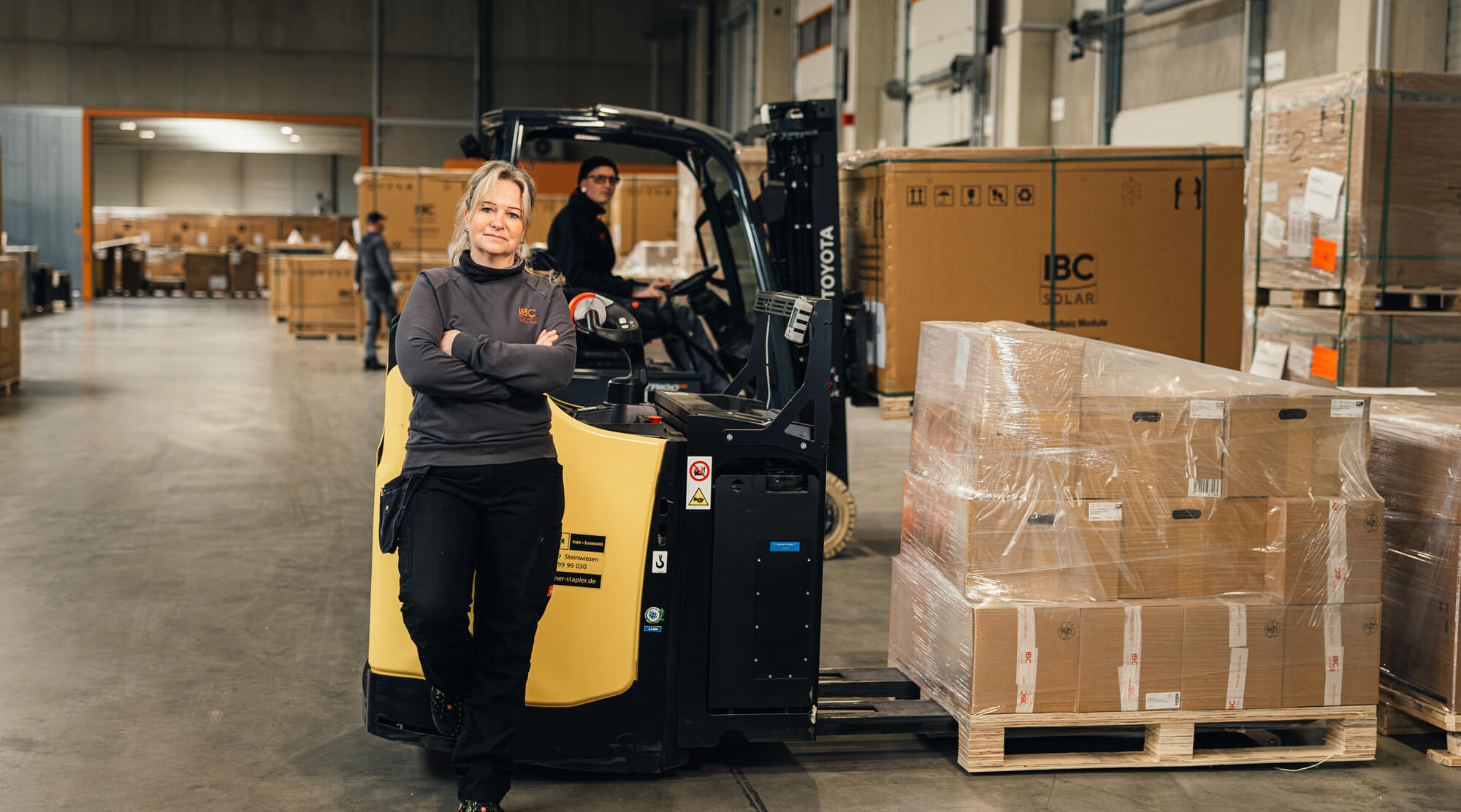 Mitarbeiter steht vor einem Gabelstapler in einer Lagerhalle, während der Stapler palettierte Kartons transportiert